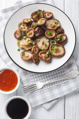 Delicious fried shiitake mushrooms with green onions close-up on a plate served with chili and soy sauces on a wooden table. Vertical top view from above