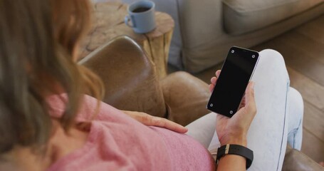 Happy caucasian pregnant woman sitting in armchair and using smartphone with copy space