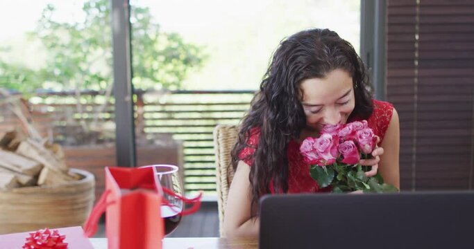 Happy biracial woman with vitiligo making valentine's day video call on laptop, smelling roses