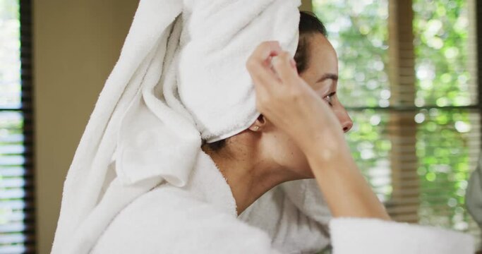 Profile of happy biracial woman with vitiligo with wet hair in towel, smiling to mirror in bathroom