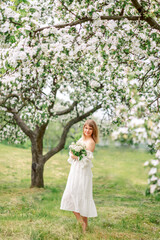 Fototapeta na wymiar young woman walks in a blooming garden in spring. A girl bride in a white dress with a bouquet in her hands