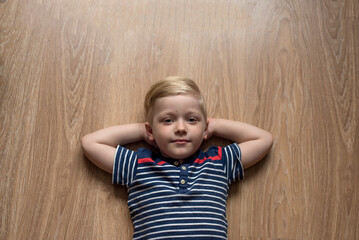 Young cute serious blond boy in stripped t-shirt is laying on the brown floor. Place for text