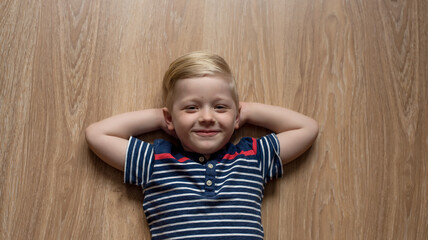Young cute happy smiled blond boy in stripped t-shirt is laying on the brown floor. Place for text