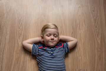 Young cute blond boy in stripped t-shirt is laying on the brown floor. Place for text