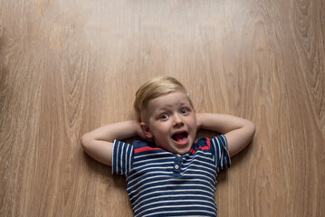 Young cute surprised blond boy in stripped t-shirt is laying on the brown floor. Place for text