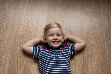 Young cute happy smiled blond boy in stripped t-shirt is laying on the brown floor. Place for text