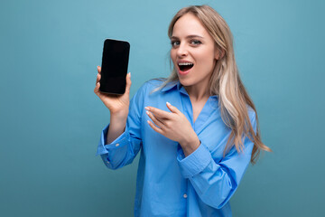 surprised blond girl holding a smartphone with a screen at the camera with an empty mockup for a website on a blue background