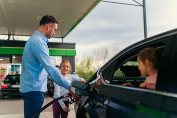 The father pours fuel into the car while his daughter watches him and keeps him company, younger...