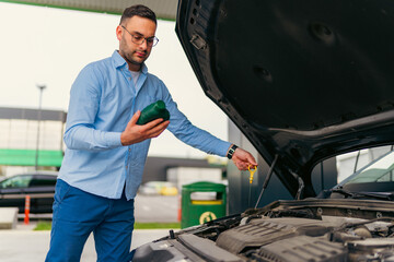 The man is standing in front of the raised hood, checking and changing the oil at the gas station