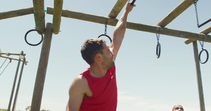 Fit caucasian man in red vest hanging from trapeze bars on obstacle course in sun