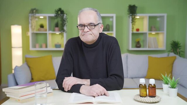 The wistful and thoughtful old man drinks water and relaxes.
The old man sitting alone at home is rueful and thoughtful, drinks the water on the table and feels relief.
