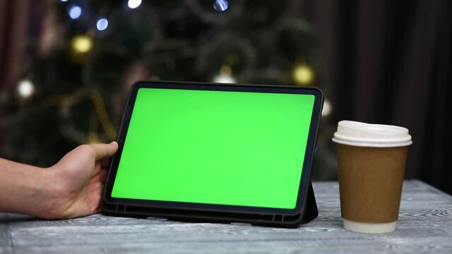 tablet with green screen in hands next to glass of coffee on christmas background