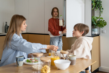 Pleased teen girl pulls bottle of milk out of fridge for cornflakes. Happy family morning table time. Smiling daughter help mother and brother prepare breakfast before school healthy eating at home
