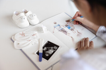 A pregnant woman is sketching the image of her family in a notebook with a pencil, baby items and...