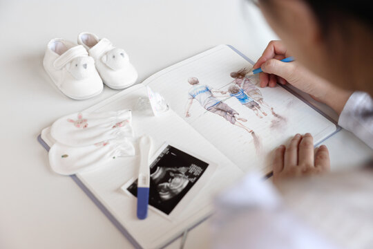 A pregnant woman is sketching the image of her family in a notebook with a pencil, baby items and ultrasound pictures are placed