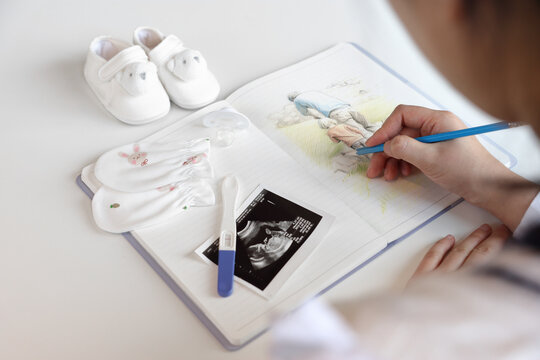 A pregnant woman is sketching the image of her family in a notebook with a pencil, baby items and ultrasound pictures are placed