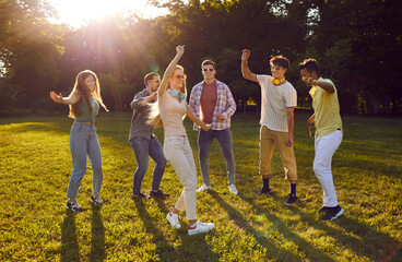 Company of young multicultural friends having fun dancing on green lawn in park on summer day. Friendship and active lifestyle concept. Guys and girls are spending free time on weekend together.