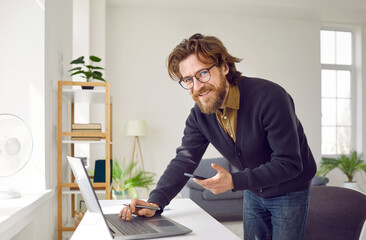 Successful satisfied Caucasian man entrepreneur uses laptop and mobile phone running own business fulfills customer orders via Internet dressed in casual clothes stands near with desk in home office