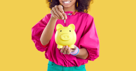 Happy kid saving up some money. Little child holding yellow piggy bank. Cheerful Afro American girl puts one coin inside her little piggybank. Cropped shot. Banner background. Finance concept