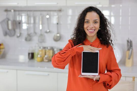 Smart and active latin woman standing while whowing tablet with happy smiling face in kitchen. Anywhere and work ifestyle with technology concept.