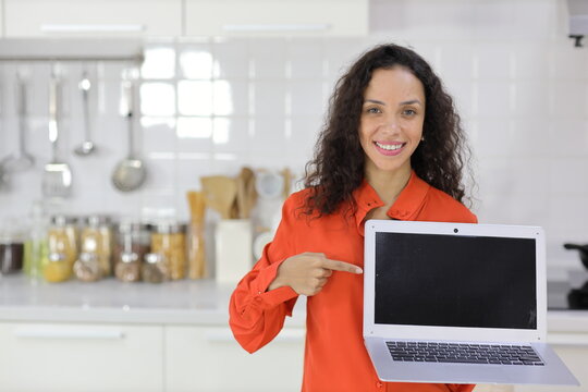 Smart and active latin woman standing while whowing and pointing at computer with happy smiling face in kitchen. Anywhere and work ifestyle with technology concept.