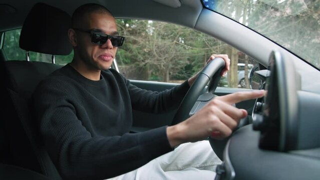 Positive smiling casually dressed young adult guy sitting on driver's seat and changing radio settings on the car cockpit. High quality 4k footage