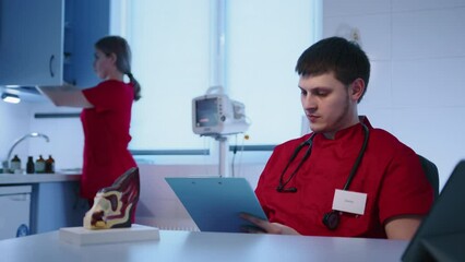 A young male veterinarian is making notes, having a stethoscope around his neck. A nurse is putting medication bottles into the cupboard. High quality 4k footage