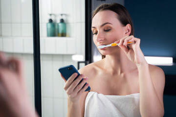 Young woman in bathroom brushing her teeth while reading messages on mobile phone. 