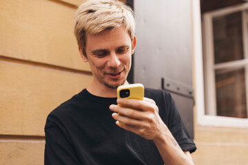 Fototapeta premium Happy hipster man walking on the street and smiling. Short-haired blonde man looking at mobile phone screen and enjoying reading messages. Man texting sms, laughing, look meme, digital communication.