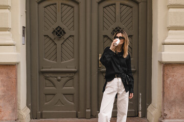 Calm stylish woman having good fashion clothes walking on street and holding tea or coffee in cup takeaway with good mood. Girl drink coffee, standing near wood green door and waiting for friends.