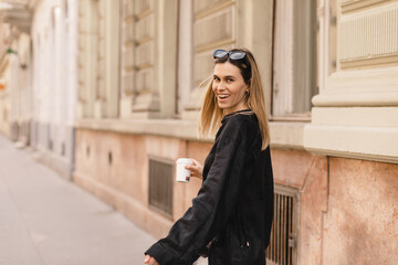 Obraz premium Smiling beautiful stylish woman having good fashion clothes walking on street and holding some coffee in cup takeaway with good summer mood. Girl look happy, turn around or spinning.