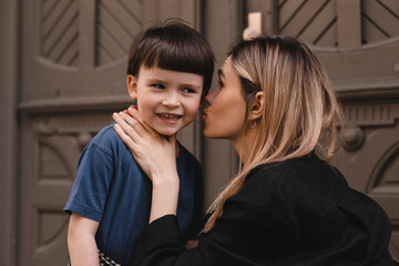 Happy mom tell secret or joke on ear her son. They look happy and laughing. Mother and children near door outside, walking on the street. Happy family, parent, childhood concept.