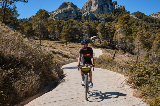 Man cyclist on a gravel bike riding on the road in the hills with a view of the mountains.Man cyclist wearing cycling kit and helmet.Gravel bike with cycling bag.Sport motivation.Alicante region,Spain