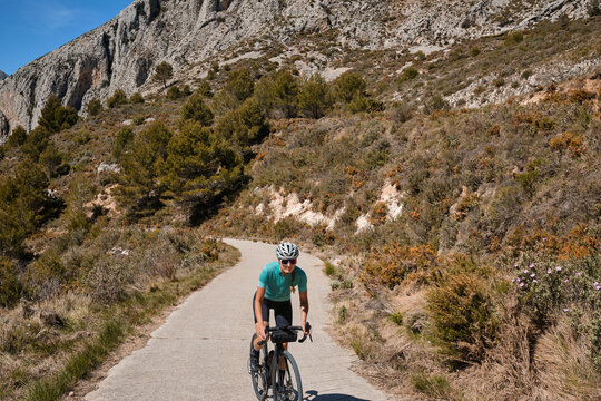 A female cyclist climbing a mountain road on a gravel bike, wearing a cycling kit and helmet.The cyclist riding between mountains, creating a beautiful and motivational image of an athlete.Spain