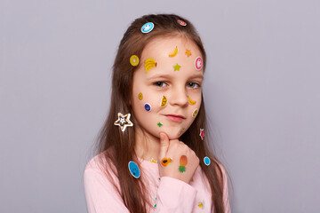 Portrait of cunning sly pensive little brown haired little girl covered with stickers posing...