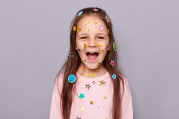 Indoor shot of excited extremely happy little brown haired little girl covered with stickers screaming with joyful expression, rejoicing posing isolated over gray background - obrazy, fototapety, plakaty