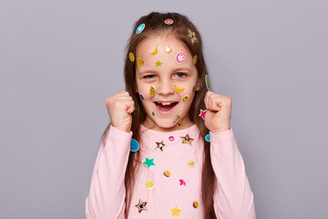 Portrait of overjoyed little brown haired little girl covered with stickers posing isolated over gray background, clenched fists, celebrating victory, screaming hurray.