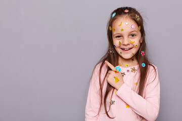 Image of happy joyful little brown haired little girl covered with stickers posing isolated over gray background, pointing finger aside at copy space for promotion.