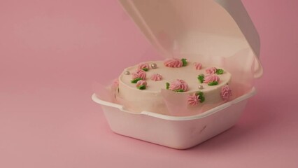 Small traditional Korean style bento cake decorated with pink whipped cream flowers. Woman hand opens white gift box with little spring cake on the pink pastel background