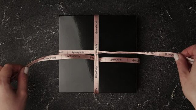 Beautiful woman hands unpack a gift. 
Female hands untie a pink bow on a black gift box. Close up. Top view. 