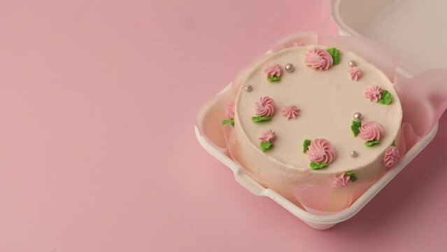 Small traditional Korean style bento cake with white cream cheese frosting decorated with pink whipped cream flowers. Little spring cake in the white gift box on the pink pastel background. Top view