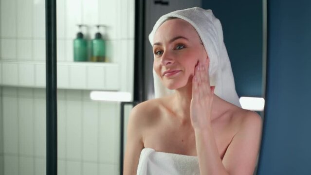 An attractive woman wearing a towel in the bathroom, applying a nourishing cream for facial skin care, looking in the mirror, performing a daily morning beauty routine