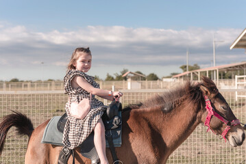 Adorable little caucasian girl in a summer dress smiling while sitting on a brown horse during the...