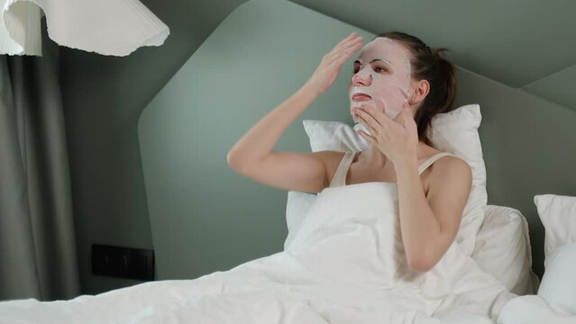 Portrait of a nice young woman lying on a bed in a bedroom with a face mask, enjoying spa treatments