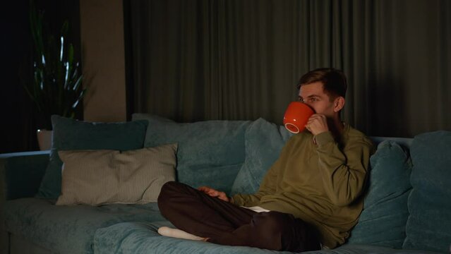 Charismatic young man watching TV. Streaming digital television ad concept. A young man watches TV and drinks tea alone.