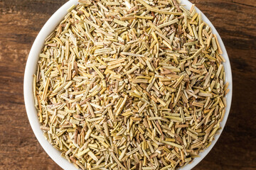 Dry rosemary leaves in bowl on rustic wooden table. Top view