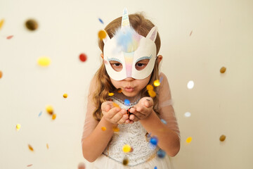  A child in a carnival mask of a unicorn, hand-made from glitter foamiran and faur fux. Little cute cheerful comical girl in the role of a unicorn, preparing for the carnival, masquerade, holidays.