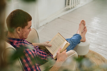 A serene man reading a captivating book, as green foliage creates a calming atmosphere at home. Man leisurely reading a book in a comfortable chair