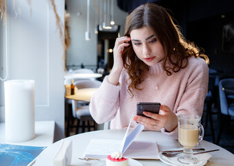 Cheerful female student with smile sitting at table in cafe reading information on mobile phone.