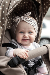 Portrait of happy smiling with blue eyes girl sitting in stroller, looking at camera. Adorable infant baby with headband on head in pram. Concept of infancy, childhood, parenthood. Copy ad text space